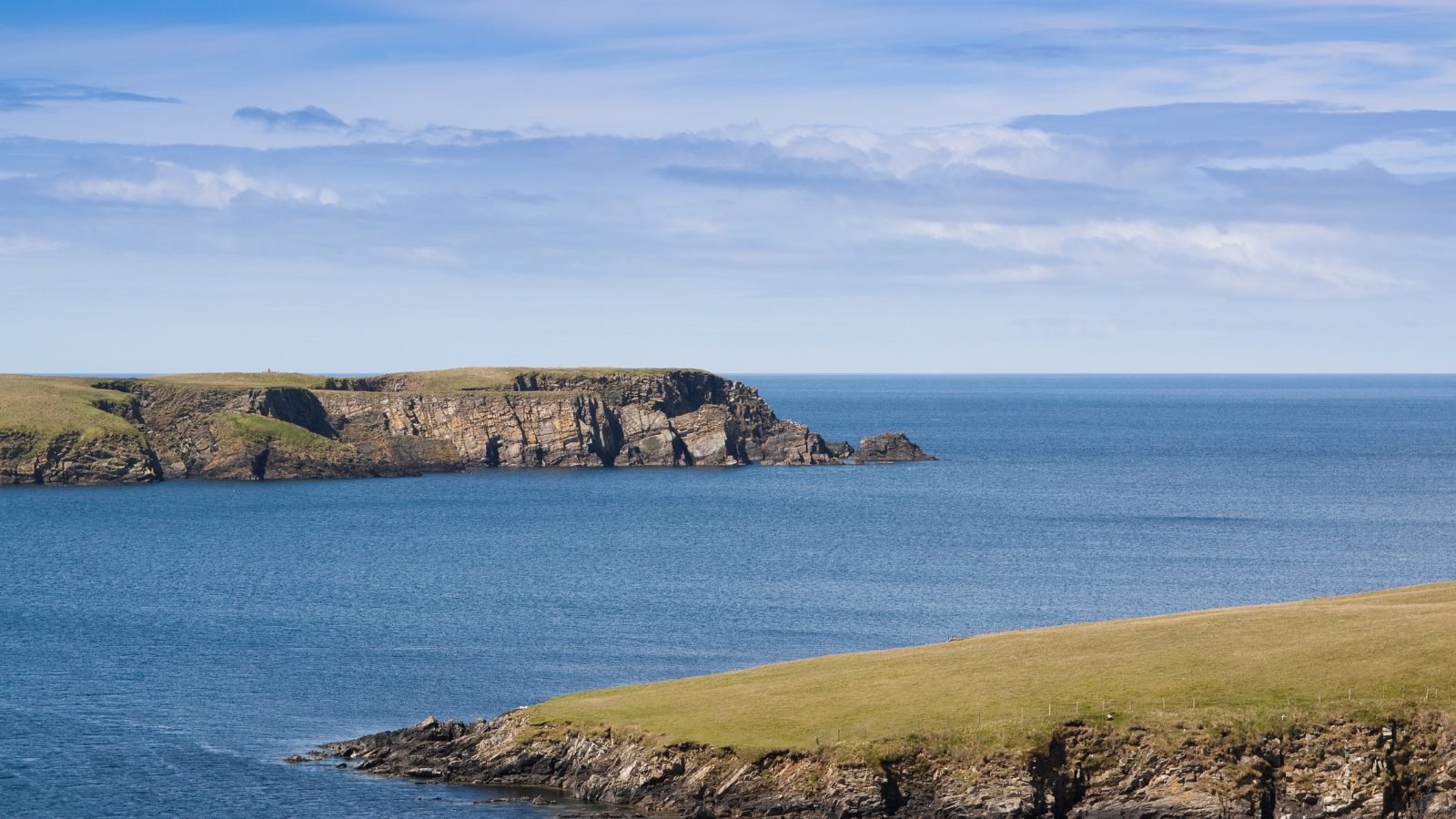 Cliffs and grassland in the Shetland Isles beside a blue sea, under a lightly clouded sky